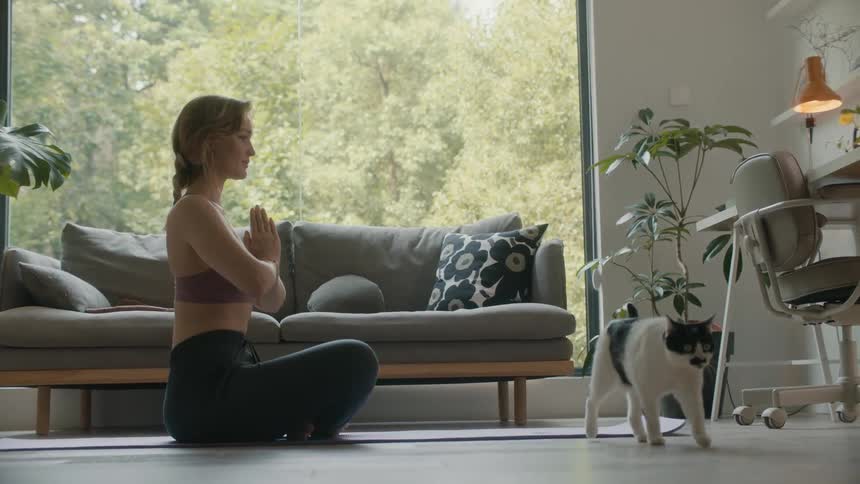 A woman is sitting on the floor practicing yoga as a black and white cat walks across the room