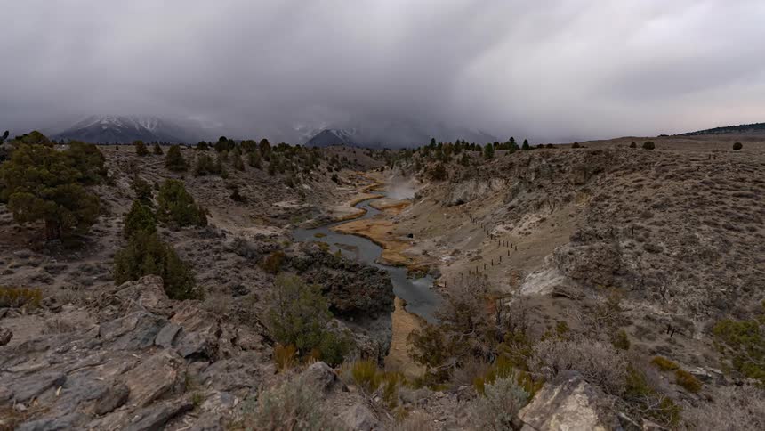 Low clouds move over a curving mountain stream with snow capped mountains behind
