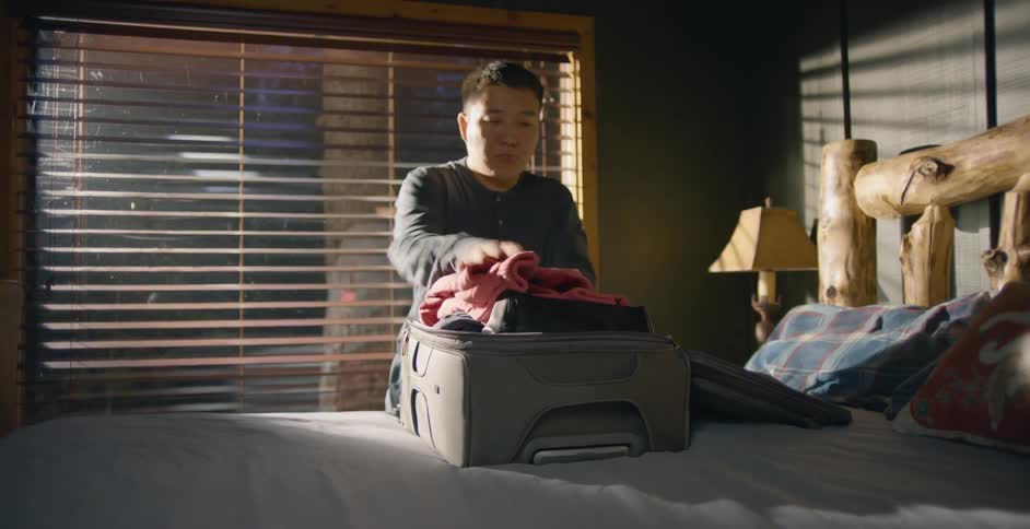 A man is putting clothes into a suitcase on a bed before zipping it shut