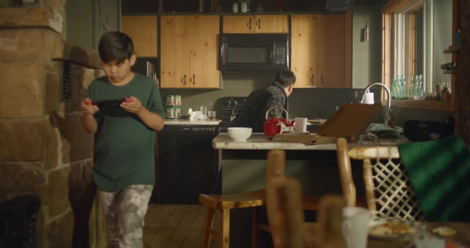 A man runs through the kitchen past his son as the boy walks and plays a video game on a portable console