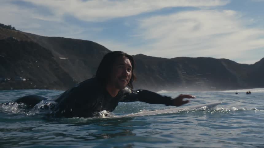 A man swims further out into the ocean on his surfboard