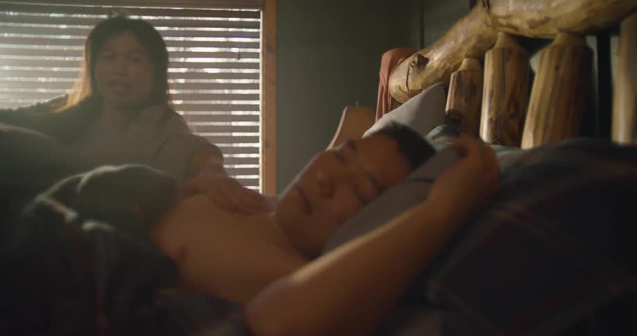 A man sleeps in bed when suddenly his wife sits up and looks at the window and then turns and wakes her husband up and she jumps out of bed as he looks at the time and jumps up