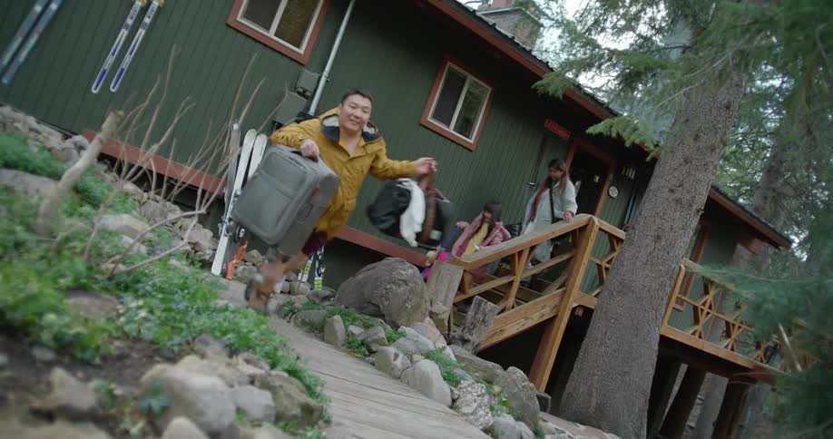 A man runs out of a cabin with suitcases as his wife and daughter trail behind him
