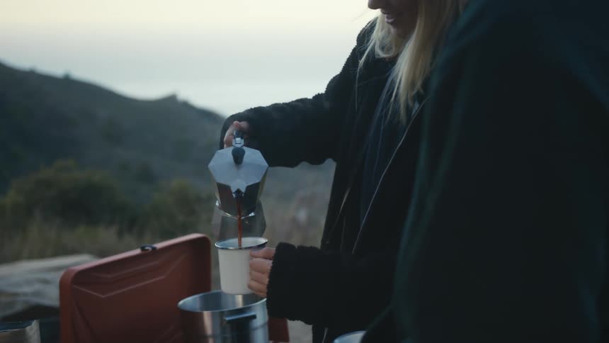 Two people are standing outside and a woman pours drinks from a metal tea pot into cups