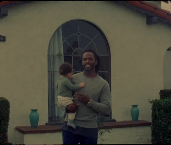 Father with braids and gray sweater holds baby boy in front of building with window