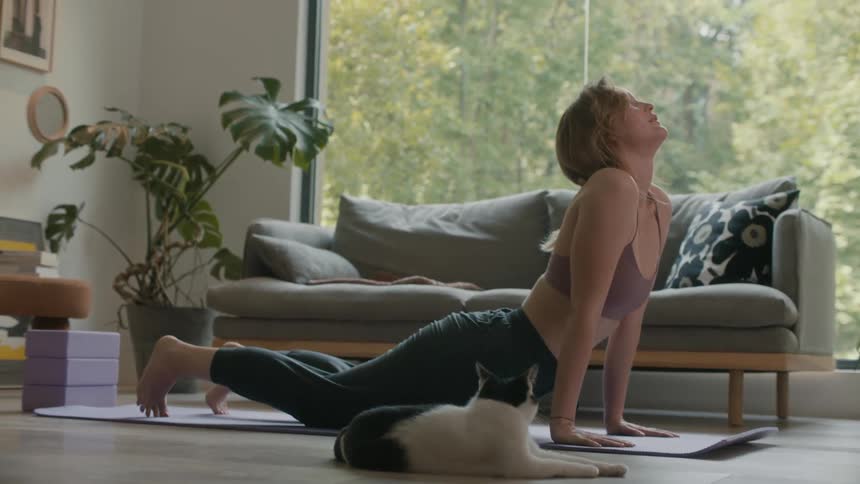 A woman is on the floor exercising on a mat and her cat is lying on the floor next to her