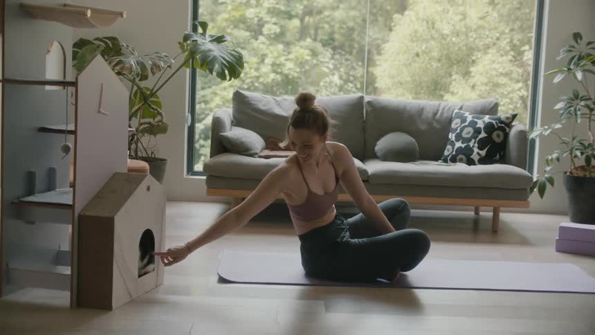 A woman is sitting on a yoga mat in her living room and reaching to pet her cat