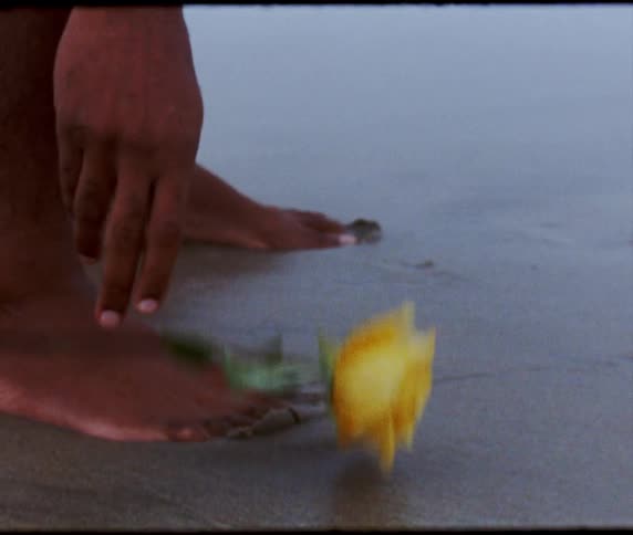 A black person places a yellow rose by their feet while standing on the beach