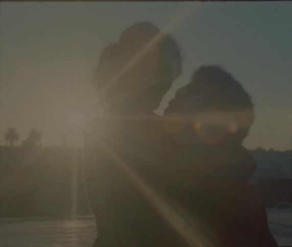 A man is hugging and lifting a woman while outside in a parking lot with a glaring sun in the background