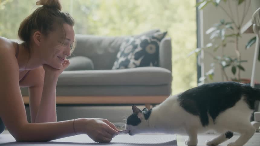 A woman is kneeling on a yoga mat in her home while holding a piece of paper that her cat is trying to grab with its mouth