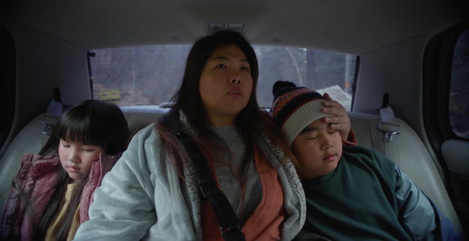A mom sitting between her two children in the backseat of a moving car hugging and comforting them as she looks anxious