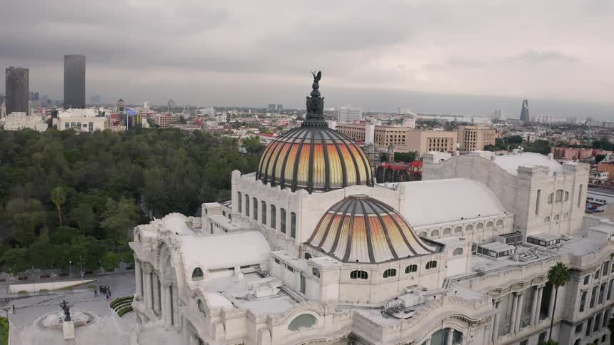 Orange and yellow colored domes top Palacio de Bellas Artes near wooded park with Mexico City in background