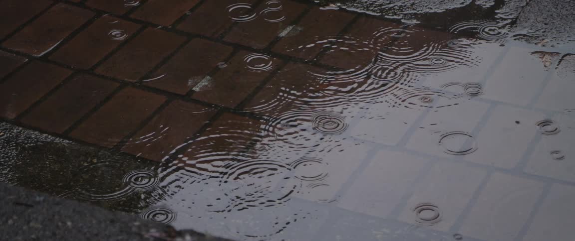 Rain drops leave ripples in a puddle on a sidewalk during the day