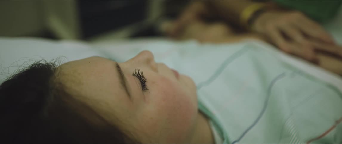 A woman is laying in a hospital bed half asleep as a loved one is touching her hand at night