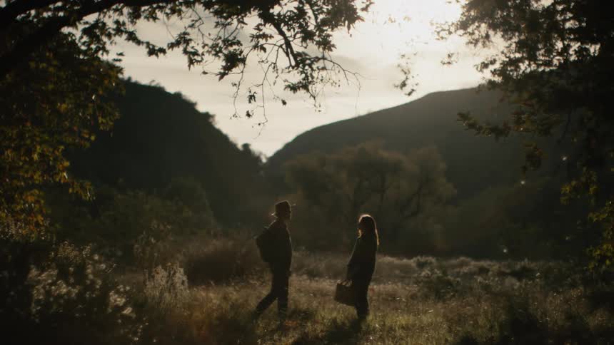 A man approaches a woman waiting for him in a clearing in the wilderness during the day