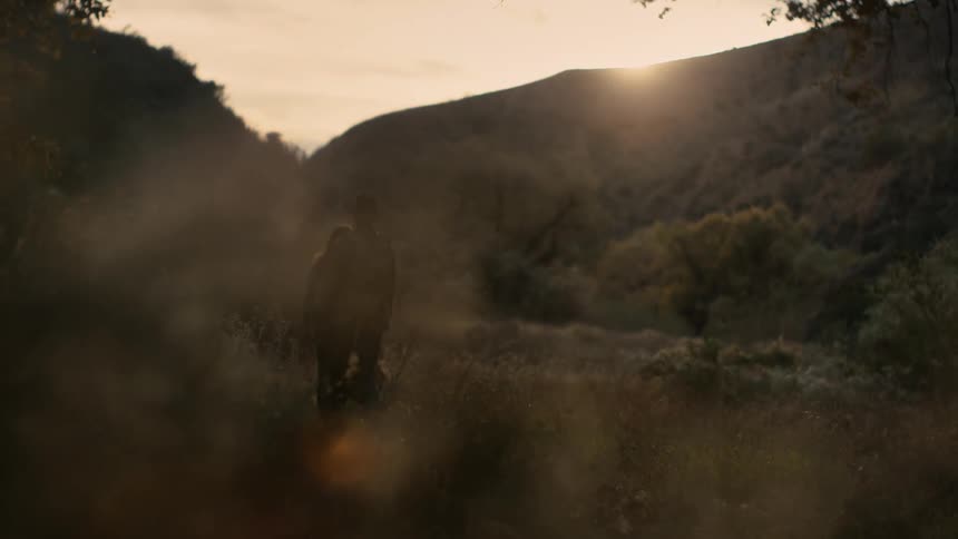 A woman carrying a picnic basket walks with a man across a grassy field surrounded by hills