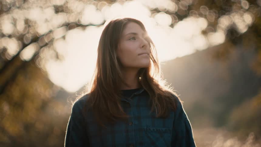A woman with long dark hair is standing outside looking at trees as the sun shines brightly behind her