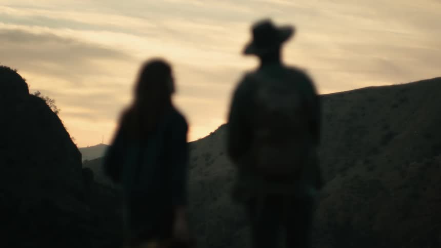 A man and woman are standing on a mountain ridge at dusk and the man is pointing out another mountain ridge