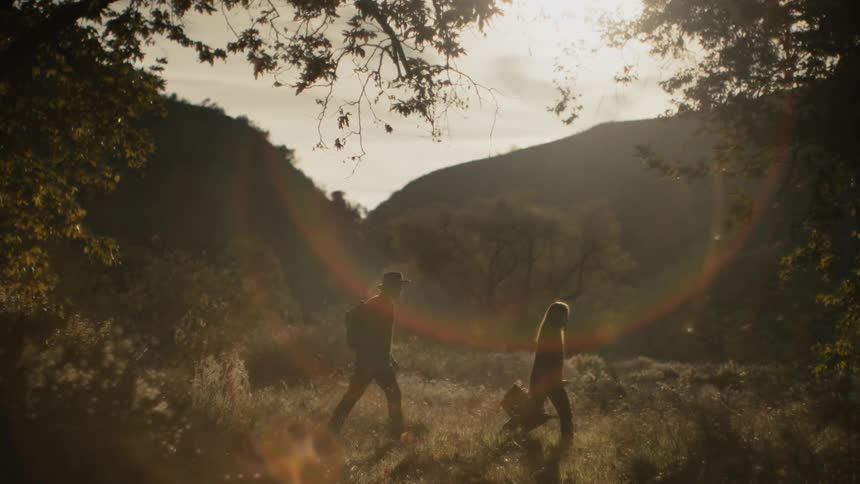 A woman leads a man through a clearing in the wilderness in the evening