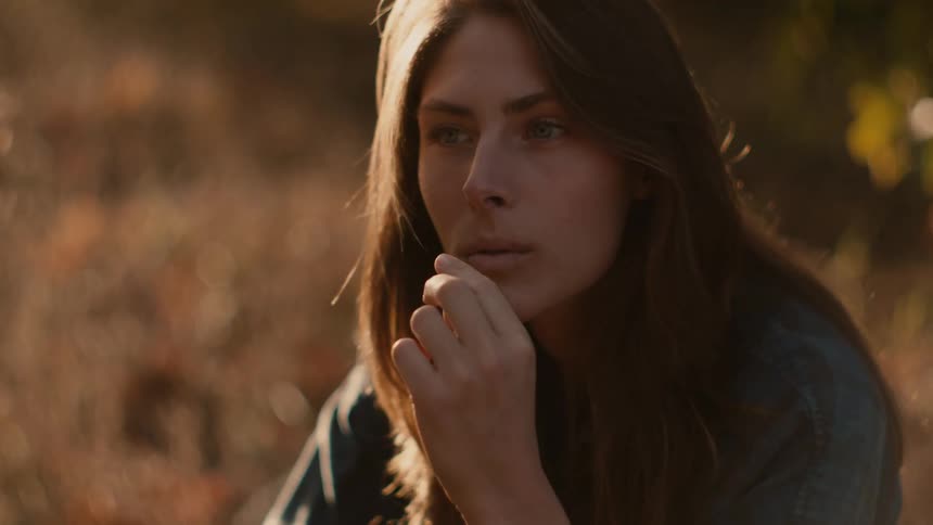 A woman with long dark hair is outside and turns her head as she puts a piece of dry grass to her mouth