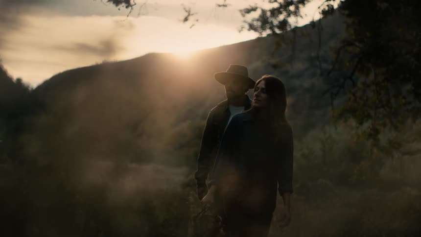 A man wearing a hat and a woman holding a picnic basket are walking towards trees as the sun sets over the mountains behind them