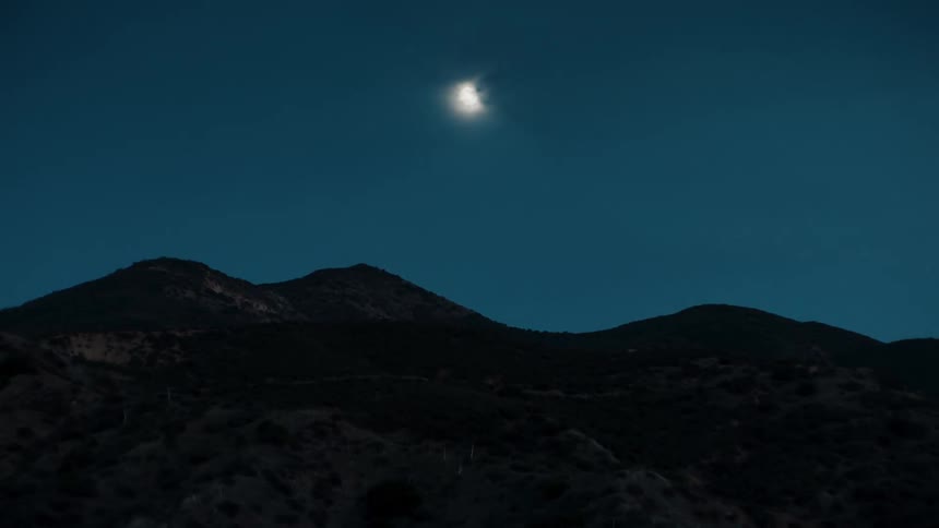A wide shot of a mountain ridge at night lit by the full moon on partly cloudy evening