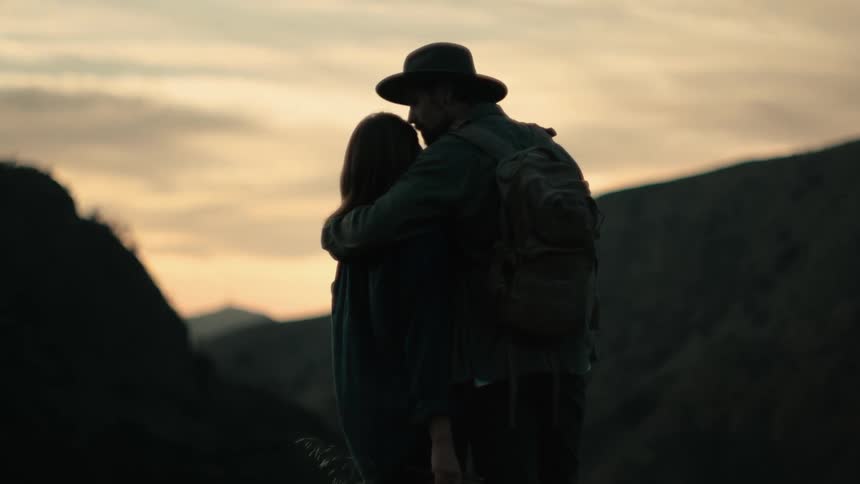 A man in a cowboy hat hugs and kisses a woman while they stand looking out at the mountains