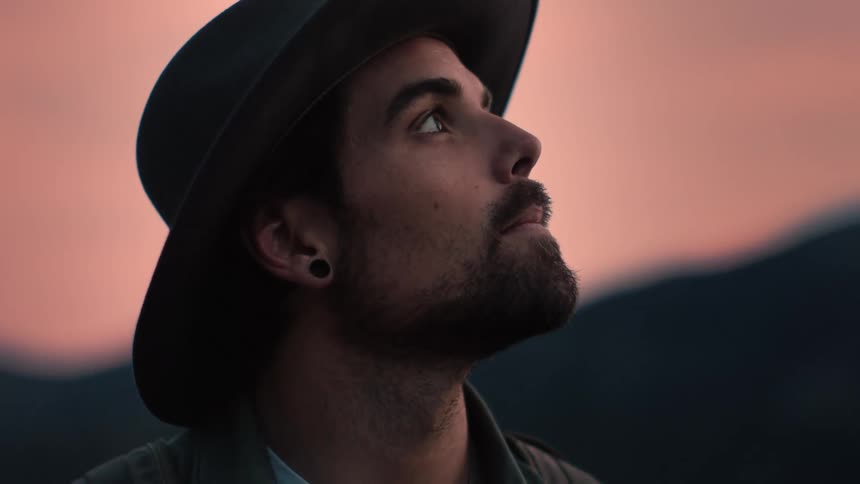 A man wearing a hat is outside looking around under a pink sky