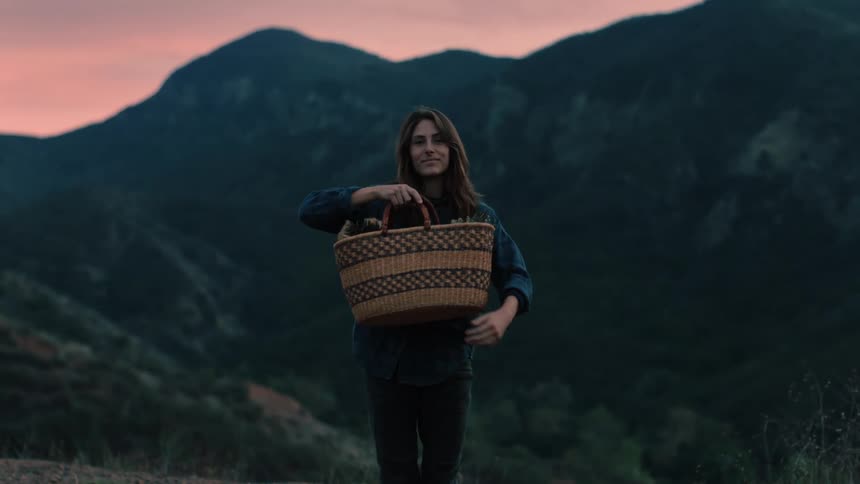 Woman standing with valley and mountains under red sky in background poses with basket that she then puts down