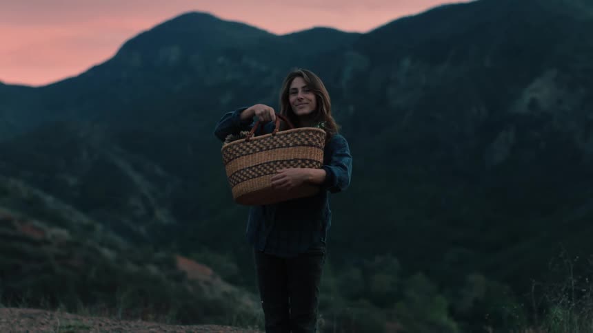 A woman picks up a basket and cradles it as she poses in front of mountains