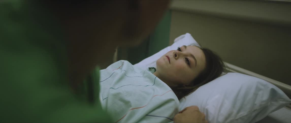 A man sits on the side of a hospital bed as a woman lies motionless in it