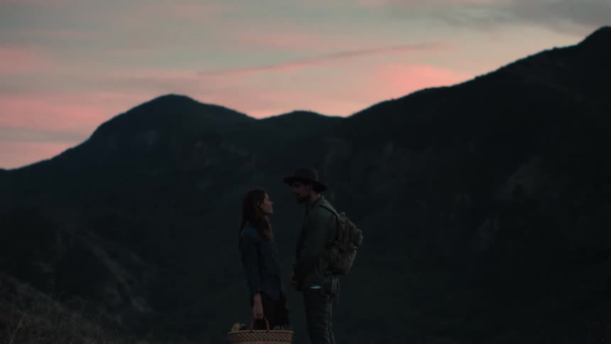 A man wearing a hat and a backpack is talking to a woman holding a picnic basket with mountains in the distance
