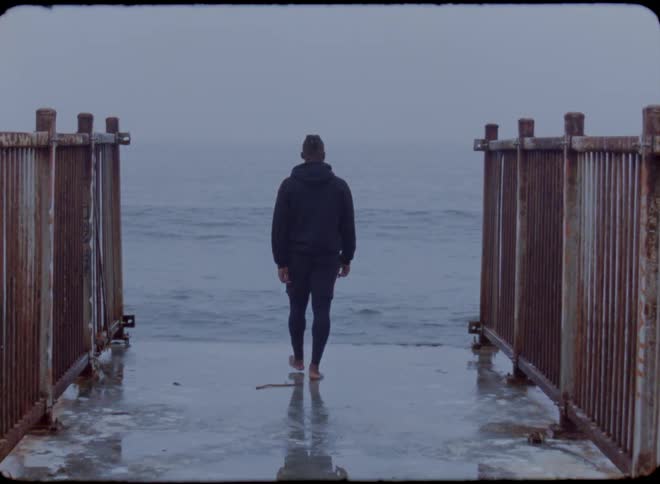 A man walks barefoot out to the edge of a pier and puts his arms out as the waves roll in