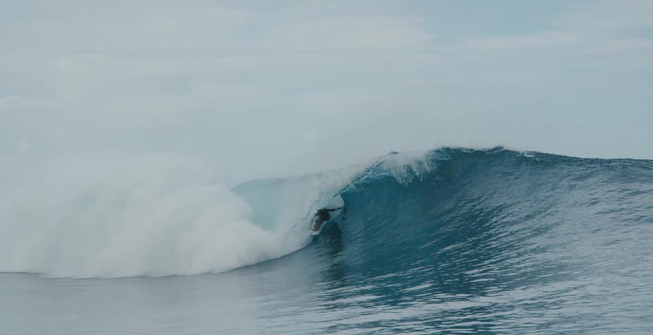 Surfer rides through barrel of crashing wave near other surfers on cloudy day