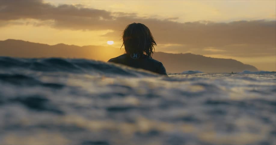 A male stands in the ocean and looks at the sunset as waves move around him