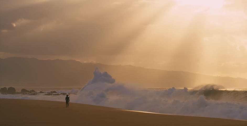 Teen surfer holding surfboard walking on the beach next to sea with crashing big waves is silhouetted against the sunset