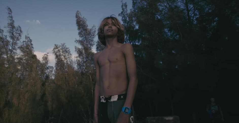 A man standing shirtless with shorts and a watch on with trees behind him