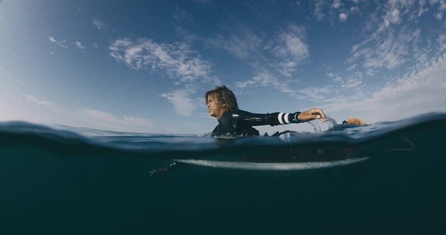 Surfer lies on surfboard and paddles it through turquoise water under a cloudy sky