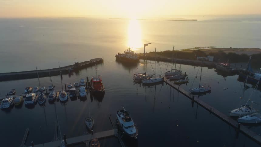 Several boats docked in a marina with a larger ship at the entrance at sunset