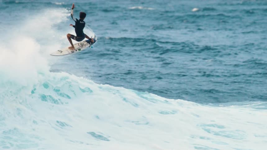 A surfer spins in the air and nearly falls off his surfboard
