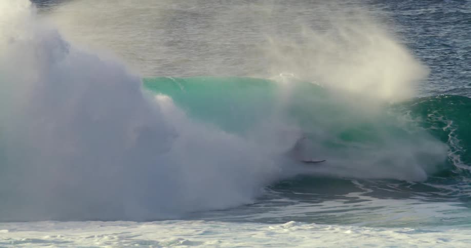A surfer stands on his surfboard and rides a big blue wave as water splashes overhead