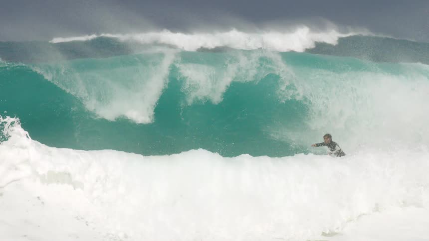 Several surfers sit on boards on one of a series of waves and then one surfer gets up on board