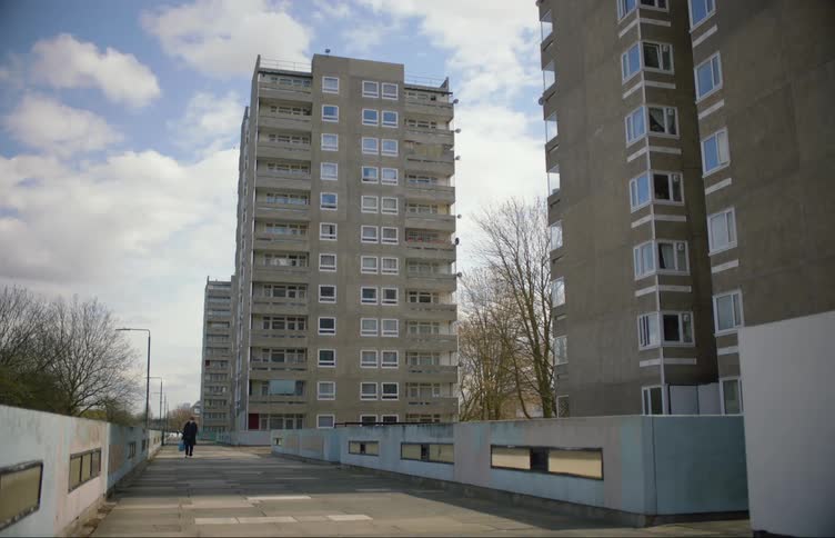 Man with mask black coat carrying blue bag walks on road by apartment buildings in the street