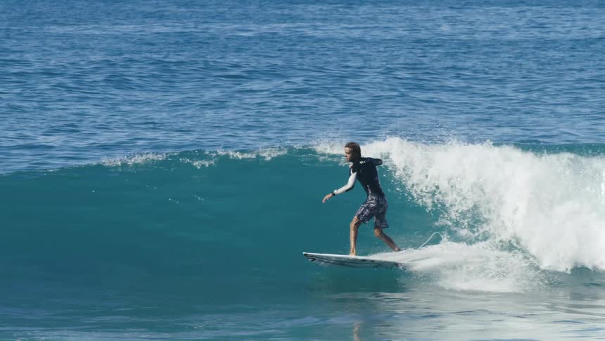 Surfer rides surfboard across low wave jumping and spinning in the air as another person floating in water watches