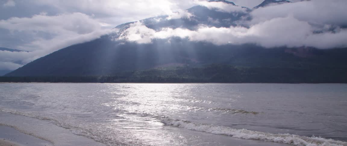 Waves roll in over a beach as the sun shines through the clouds above mountains