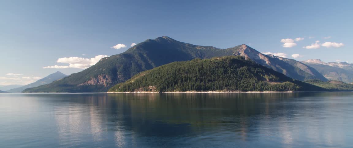 Water flows along shore reflecting forest covered mountain under cloudy sky
