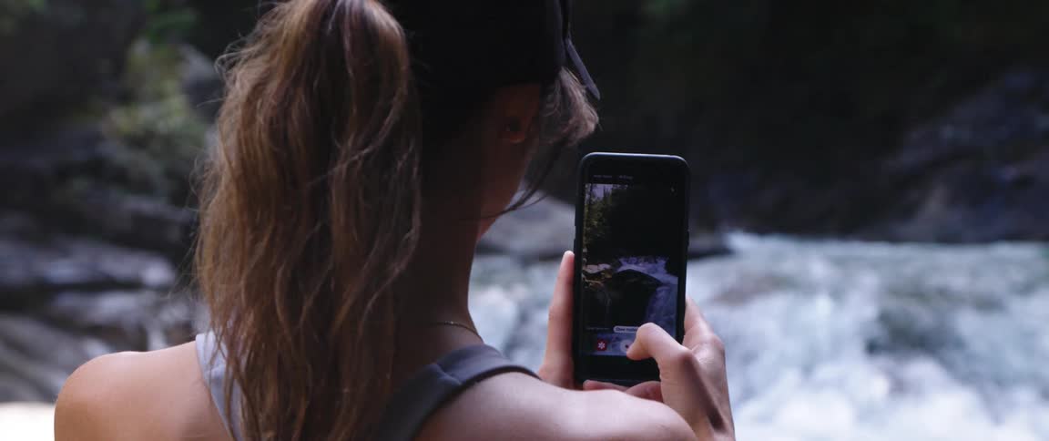 Woman with brown wavy hair and sports bra fims recors waterfall cascading down on smartphone in the forest