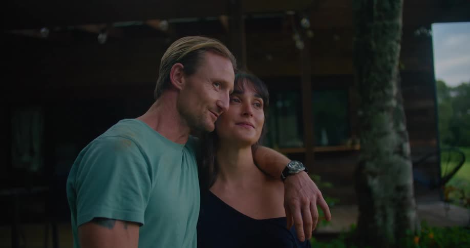 Man with turquoise t shirt and woman with brown wavy hair and black dress hold each other pointing and looking over green field next to countryhouse in the countryside at dusk