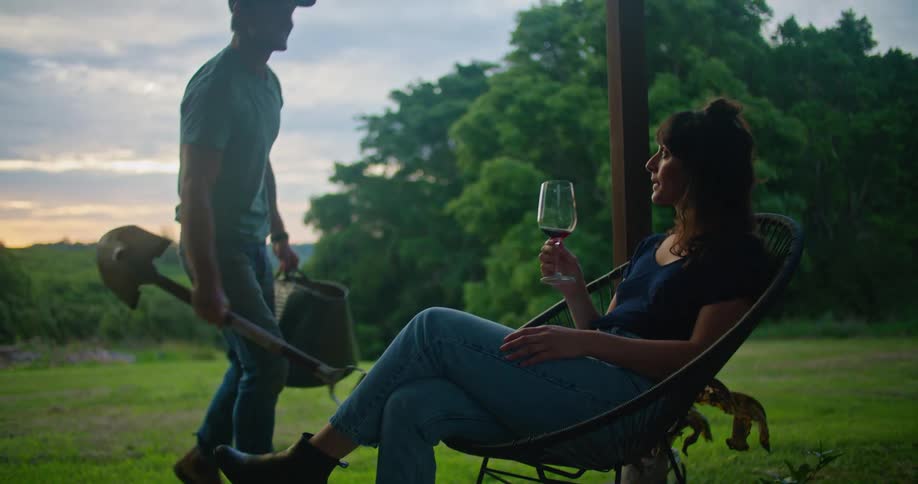 A man walks up to a woman sitting on a porch drinking a glass of wine and bends down to kiss her
