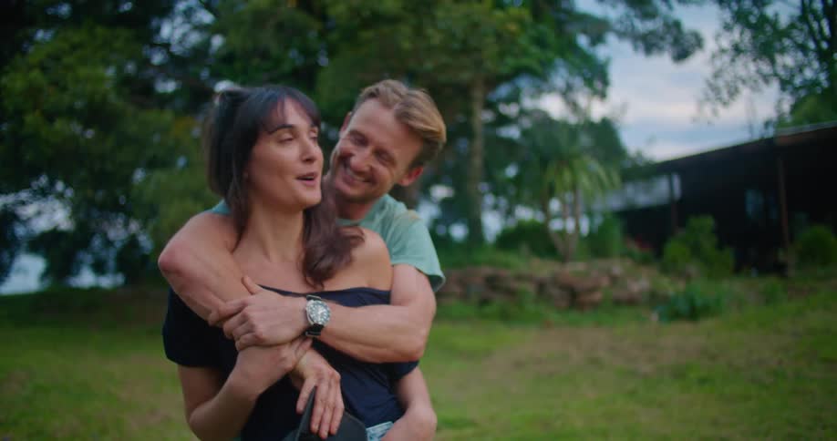 Man stands hugging woman from behind as they talk in large yard near their home